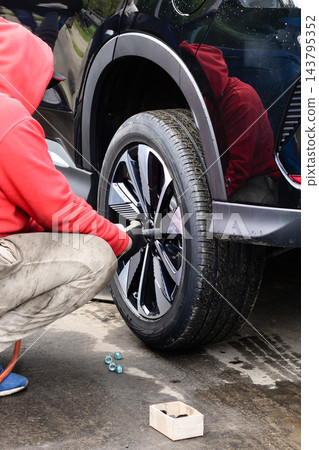 Guy changes wheel on vehicle in a parking area during the day while wearing a red hoodie Guy changes wheel on vehicle in a parking area during the day while wearing a red hoodie 143795352