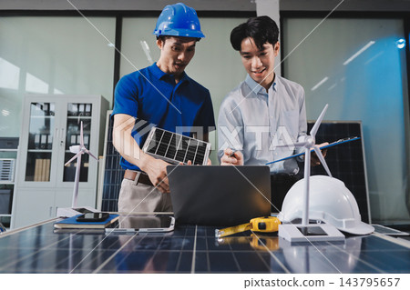 Two Asian engineers wearing hardhats and safety vests discussing solar panel installation and renewable energy project in the office. 143795657