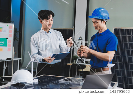 Two Asian engineers wearing hardhats and safety vests discussing solar panel installation and renewable energy project in the office. 143795663