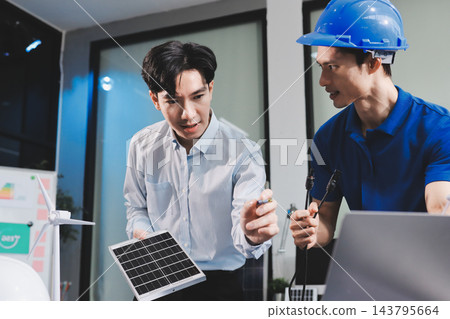 Two Asian engineers wearing hardhats and safety vests discussing solar panel installation and renewable energy project in the office. Two Asian engineers wearing hardhats and safety vests discussing solar panel installation and renewable energy project in the office. 143795664