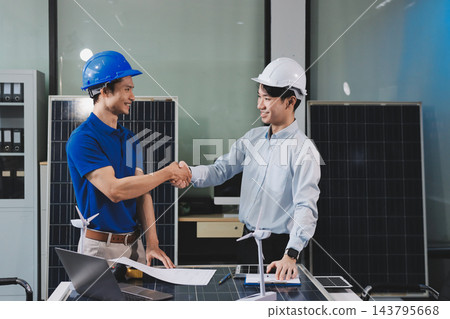 Two Asian engineers wearing hardhats and safety vests discussing solar panel installation and renewable energy project in the office. 143795668