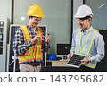 Two Asian engineers wearing hardhats and safety vests discussing solar panel installation and renewable energy project in the office. 143795682