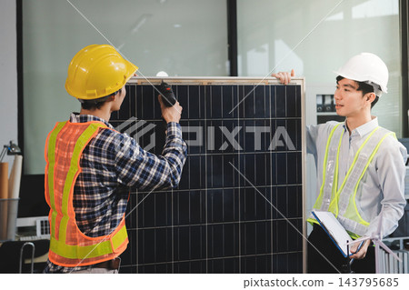 Two Asian engineers wearing hardhats and safety vests discussing solar panel installation and renewable energy project in the office. 143795685