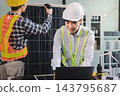 Two Asian engineers wearing hardhats and safety vests discussing solar panel installation and renewable energy project in the office. 143795687