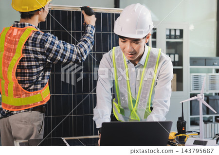 Two Asian engineers wearing hardhats and safety vests discussing solar panel installation and renewable energy project in the office. 143795687