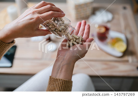 Woman pouring dietary supplement capsule pills from bottle into her hand, daily wellness routine, nutrition, healthy lifestyle and supplement use 143797269