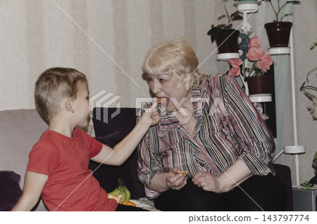 An elderly woman a grandmother brings fruit on a plate to her young grandson in the room and feeds him. Caring for children and the elderly parental love. An elderly woman a grandmother brings fruit on a plate to her young grandson in the room and feeds him. Caring for children and the elderly parental love. 143797774