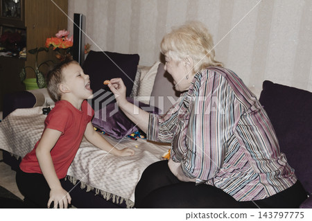 An elderly woman a grandmother brings fruit on a plate to her young grandson in the room and feeds him. Caring for children and the elderly parental love. 143797775