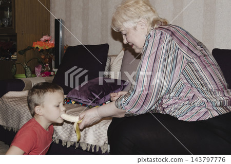 An elderly woman a grandmother brings fruit on a plate to her young grandson in the room and feeds him. Caring for children and the elderly parental love. 143797776