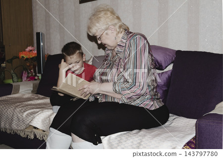 grandmother is reading colorful book grandson, has leaned in closer to listen. grandmother wearing glasses, boy, dressed red T-shirt, is intently examining the book and listening to the story. 143797780