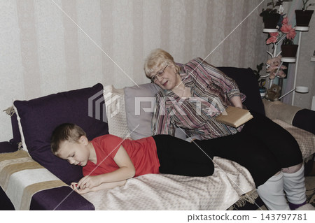 grandmother is reading colorful book grandson, has leaned in closer to listen. grandmother wearing glasses, boy, dressed red T-shirt, is intently examining the book and listening to the story. grandmother is reading colorful book grandson, has leaned in closer to listen. grandmother wearing glasses, boy, dressed red T-shirt, is intently examining the book and listening to the story. 143797781