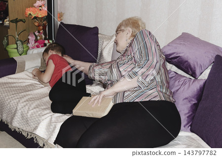 grandmother is reading colorful book grandson, has leaned in closer to listen. grandmother wearing glasses, boy, dressed red T-shirt, is intently examining the book and listening to the story. grandmother is reading colorful book grandson, has leaned in closer to listen. grandmother wearing glasses, boy, dressed red T-shirt, is intently examining the book and listening to the story. 143797782