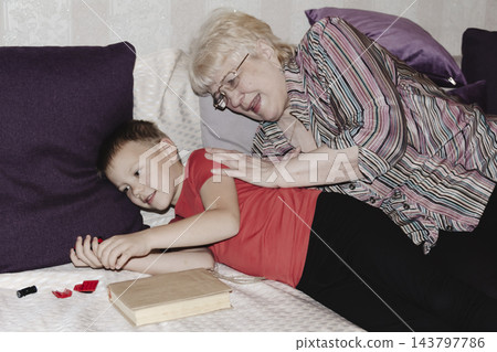 grandmother is reading colorful book grandson, has leaned in closer to listen. grandmother wearing glasses, boy, dressed red T-shirt, is intently examining the book and listening to the story. grandmother is reading colorful book grandson, has leaned in closer to listen. grandmother wearing glasses, boy, dressed red T-shirt, is intently examining the book and listening to the story. 143797786