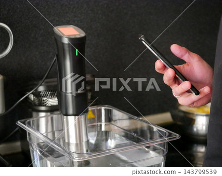 Man using smartphone to control sous vide immersion circulator in clear water container. Innovative kitchen appliance for precision cooking and professional culinary technique at home. Man using smartphone to control sous vide immersion circulator in clear water container. Innovative kitchen appliance for precision cooking and professional culinary technique at home. 143799539