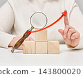 Woman's hand pointing at a red growth arrow above a magnifying glass and wooden cubes 143800020