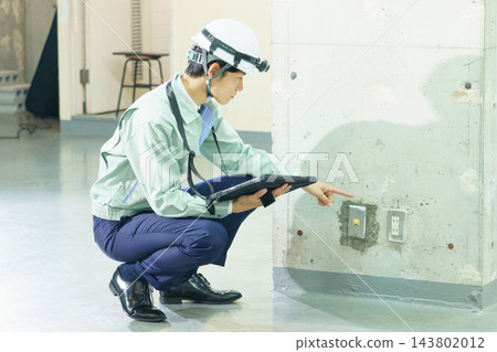 A young Japanese worker inspects electrical equipment on the wall of a construction site. 143802012