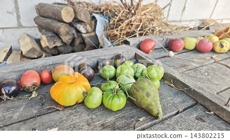 Assorted autumn vegetables and fruit arranged on a weathered wooden farm table with stacked firewood and straw in a rustic outdoor still life 143802215