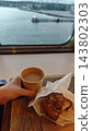 A hand holds coffee beside a glazed pastry on a wooden table, with calm water, a dock, and distant shoreline visible through the ferry window 143802303