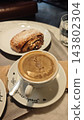 Coffee with a cinnamon pastry on a ceramic plate, spoon, water glass, and handwritten lettering on a cafe table, close up of a breakfast setting 143802304