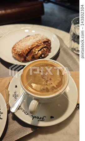 Coffee with a cinnamon pastry on a ceramic plate, spoon, water glass, and handwritten lettering on a cafe table, close up of a breakfast setting 143802304