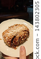 Cinnamon bun covered with pearl sugar and cinnamon on a white scalloped plate, held by a hand in a dim indoor setting 143802305