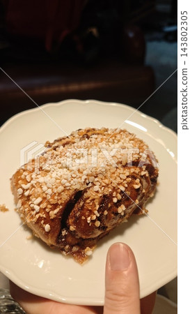 Cinnamon bun covered with pearl sugar and cinnamon on a white scalloped plate, held by a hand in a dim indoor setting Cinnamon bun covered with pearl sugar and cinnamon on a white scalloped plate, held by a hand in a dim indoor setting 143802305