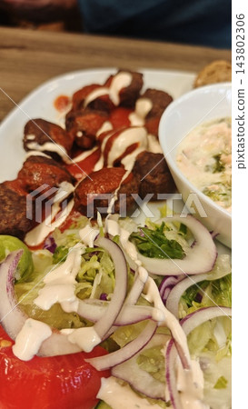 Meatballs with creamy sauce, salad, tomato, and dip arranged on a white plate over a wooden table, close up of a plated meal 143802306
