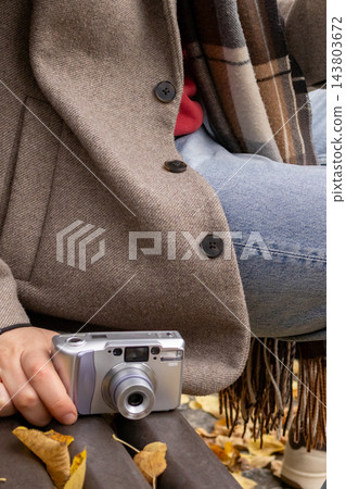 Hands of a man hold a silver compact film camera while he sits on a park bench in a beige coat and jeans with yellow leaves around. Concept of nostalgia and the revival of analog photography. Hands of a man hold a silver compact film camera while he sits on a park bench in a beige coat and jeans with yellow leaves around. Concept of nostalgia and the revival of analog photography. 143803672