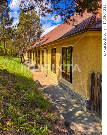Yellow house with a tiled roof stands next to a grassy slope and tall pine trees Yellow house with a tiled roof stands next to a grassy slope and tall pine trees 143804358