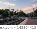 A Winter Park railroad crossing and train tracks sit under a pastel sunset sky. The quiet station area includes safety gates, rail signals, trees, fences, and brick pavement. 143804413