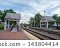 A Winter Park train station shows railroad tracks, platforms, and white covered shelters under a cloudy blue sky. The transportation scene includes brick paving, safety railings, trees, and an empty 143804414