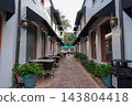 An empty cafe courtyard has brick pavement, outdoor tables, potted flowers, white buildings, and black awnings. The narrow walkway, lanterns, blue umbrellas, and shop windows create a quiet shopping 143804418