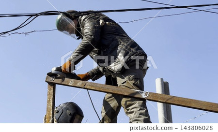 Young mechanic in protective gloves polishing iron using electric grinder wheel at construction. Professional repairman grinding metal details with circular saw on house at sunny day. Slow mo 143806028