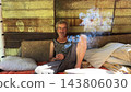 Portrait of homeless adult man sitting on worn couch placed and smoking outdoor. The setting underscores neglect and impermanence, presenting a raw and unfiltered glimpse of difficult living situation 143806030