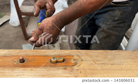 Adult male hands use a screwdriver to remove a door lock mechanism from a wooden door outdoor. Worker carefully loosens the screws and disassembles the hardware during home repair and maintenance work Adult male hands use a screwdriver to remove a door lock mechanism from a wooden door outdoor. Worker carefully loosens the screws and disassembles the hardware during home repair and maintenance work 143806031