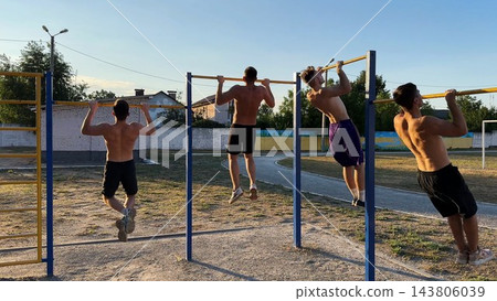 Group of strong sporty men doing pull ups on horizontal bar at sports ground. Young shirtless sportsmen exercising at nature. Muscular athletes training outdoor. Healthy and active lifestyle concept Group of strong sporty men doing pull ups on horizontal bar at sports ground. Young shirtless sportsmen exercising at nature. Muscular athletes training outdoor. Healthy and active lifestyle concept 143806039