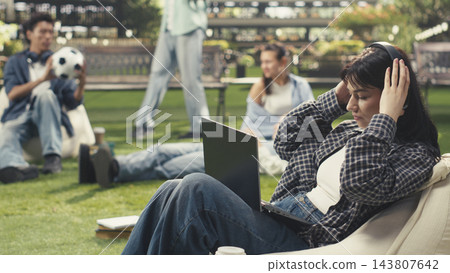 Headphones, laptop and student as woman adjusts earphones while reclining on beanbag at campus lawn. Independent study, concentration and digital learning suit personal academic routines outdoors. Headphones, laptop and student as woman adjusts earphones while reclining on beanbag at campus lawn. Independent study, concentration and digital learning suit personal academic routines outdoors. 143807642