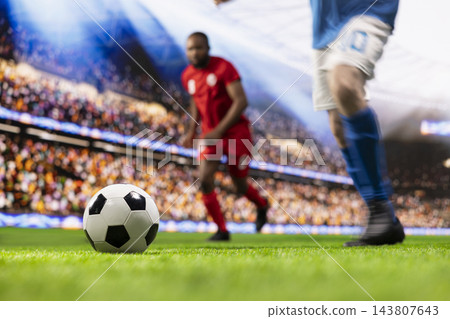 Soccer ball, grass pitch and opposing players as close-up football lies in sharp focus with blurred athletes moving behind. Competition, anticipation and match action set stadium gameplay context. 143807643