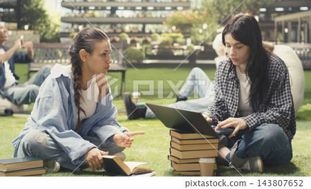 Students, laptop and books as classmates discuss coursework while studying together on grass beside stacked textbooks. Collaboration, peer learning and teamwork support academic study on campus. Students, laptop and books as classmates discuss coursework while studying together on grass beside stacked textbooks. Collaboration, peer learning and teamwork support academic study on campus. 143807652
