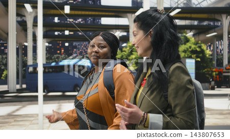 Travel companions, conversation and bus terminal as two women walk together talking beside parked coaches. Social connection, mobility and passenger experience frame shared journeys through station. Travel companions, conversation and bus terminal as two women walk together talking beside parked coaches. Social connection, mobility and passenger experience frame shared journeys through station. 143807653