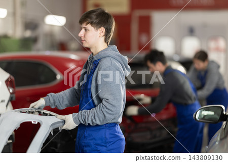 Young auto mechanic carries freshly painted bumper in his hands to be installed on car Young auto mechanic carries freshly painted bumper in his hands to be installed on car 143809130