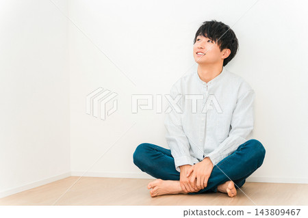 一位二十多歲的年輕男子盤腿坐在房子的地板上，帶著微笑，抬頭望向前方。 143809467