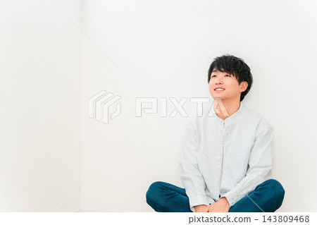 一位二十多歲的年輕男子盤腿坐在房子的地板上，帶著微笑，抬頭望向前方。 143809468