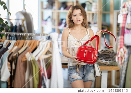 Positive young girl choosing handbag in clothing store Positive young girl choosing handbag in clothing store 143809543