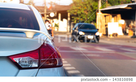 Rear view of a white car stopped on a city street during the day. Showing its detailed tail light and distant traffic on the road. Car is parked right on the red line which marks pedestrian crossing. Rear view of a white car stopped on a city street during the day. Showing its detailed tail light and distant traffic on the road. Car is parked right on the red line which marks pedestrian crossing. 143810898