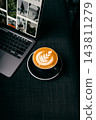 Hot coffee latte art in cup on saucer beside laptop on dark table 143811279