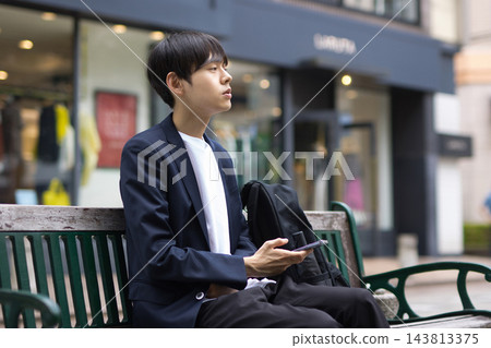 一位二十多歲的男性商人坐在長椅上看著他的智慧型手機 一位二十多歲的男性商人坐在長椅上看著他的智慧型手機 143813375