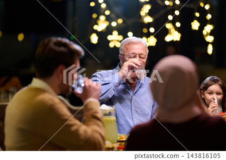 Multi-Generational Family Dinner With Grandfather Drinking Water Under Festive Lights Multi-Generational Family Dinner With Grandfather Drinking Water Under Festive Lights 143816105
