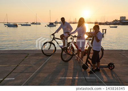 Family Cycling at Sunset on a Seaside Promenade With Electric Scooter and Boats in Harbor 143816345