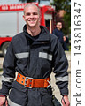 Smiling Firefighter in Protective Gear With Bright Safety Belt Standing by Fire Truck 143816347